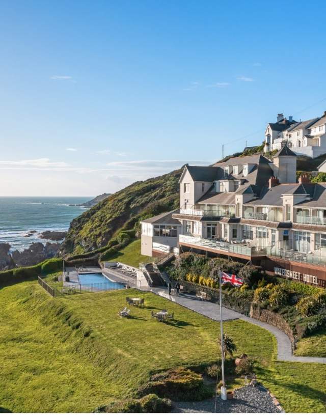 Coastal hotel on a sunlit cliff with a pool and gardens, overlooking a beach and ocean. British flag in foreground, houses atop the hill, clear skies.