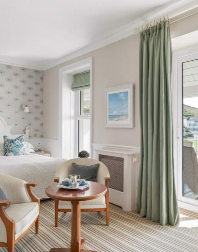 Bright hotel room with a cosy double bed, patterned wallpaper, and a small table set with tea. Glass doors open to a scenic view of hills and houses.