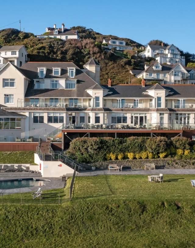 Seaside hotel on a hillside with large windows and balconies, overlooking a pool and lawn. Bright, sunny day with blue sky, evoking a tranquil, inviting mood.