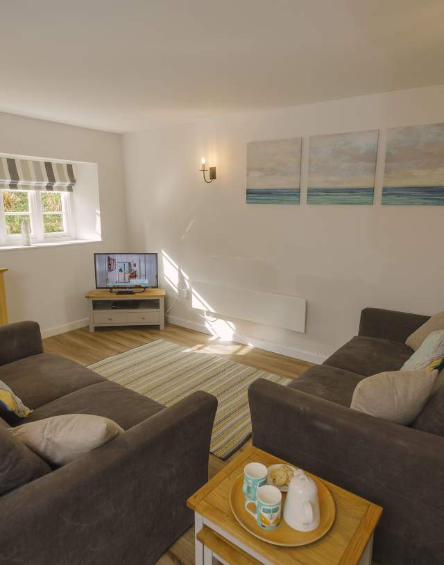 Cosy living room with two brown sofas facing a TV on a wooden stand. Soft lighting, a striped rug, and ocean-themed wall art add warmth and tranquility.