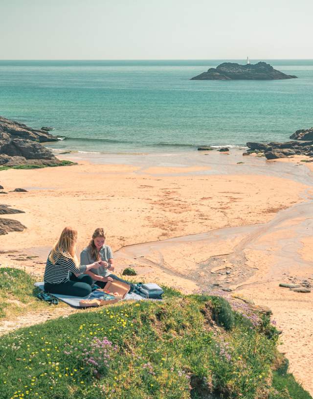 Two women sitting on a sandy beach, enjoying the sun and ocean view, with gentle waves in the background.