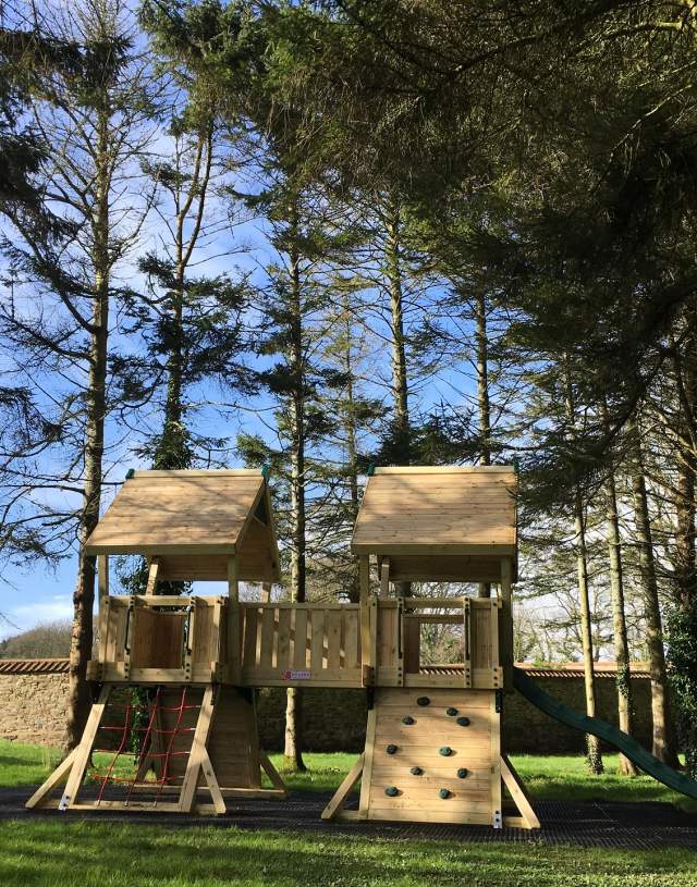 Wooden playground set with a slide, climbing wall, and rope ladder amidst tall trees under a clear blue sky, creating a serene outdoor scene.