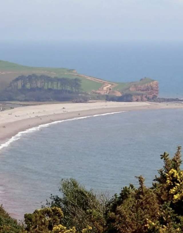 Aerial view of a broad, curved beach bordered by calm, blue sea and green, shrub-covered cliffs. The scene conveys tranquility and natural beauty.
