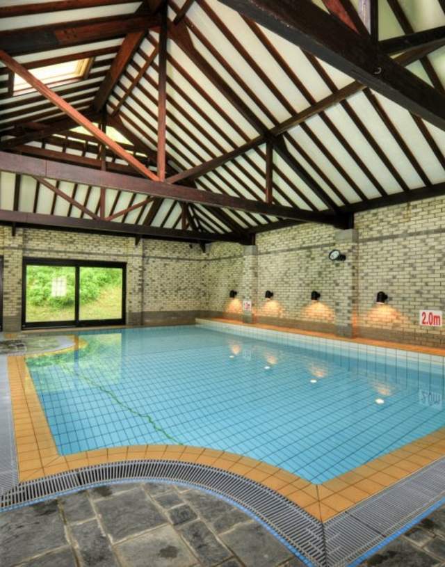 Indoor pool with rustic wooden beams and brick walls, sunlit through large windows. The pool is calm, with seating nearby, creating a serene atmosphere.
