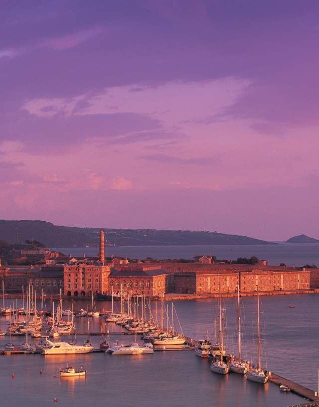 Harbour scene at sunset with pink and purple sky, featuring a marina full of sailboats. An old fort structure rests on the calm water's edge.