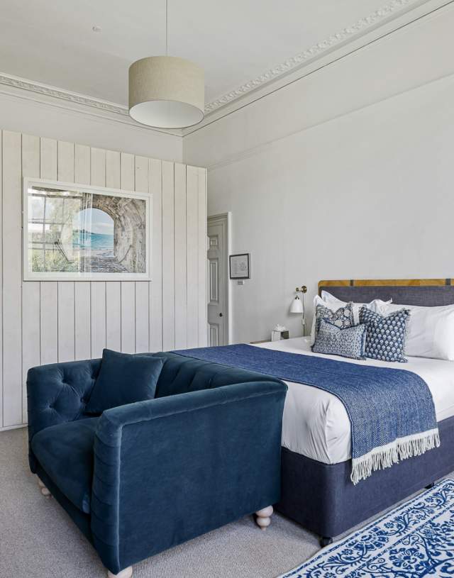 A bright, stylish bedroom with a plush navy blue sofa, a large bed adorned with blue and white accents, and a patterned rug. There's a small wooden side table and minimal decor, including a wall mirror above a corner basin. The room exudes a cosy and elegant atmosphere.