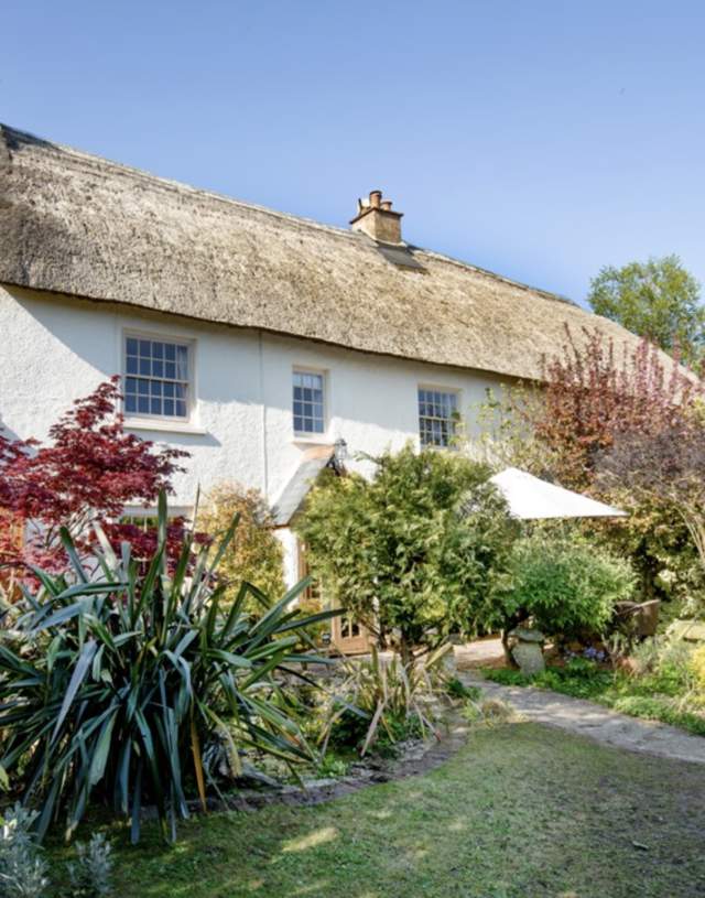 Thatch-roofed cottage with white walls, surrounded by a lush, colourful garden with various plants. A peaceful, sunny day, exuding charm and tranquillity.