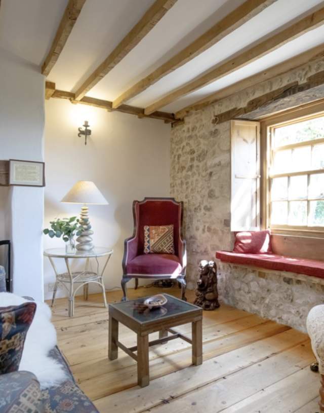 Cosy rustic living room with exposed beams, stone walls, a window seat, and vintage furniture. Warm tones and natural light create an inviting atmosphere.