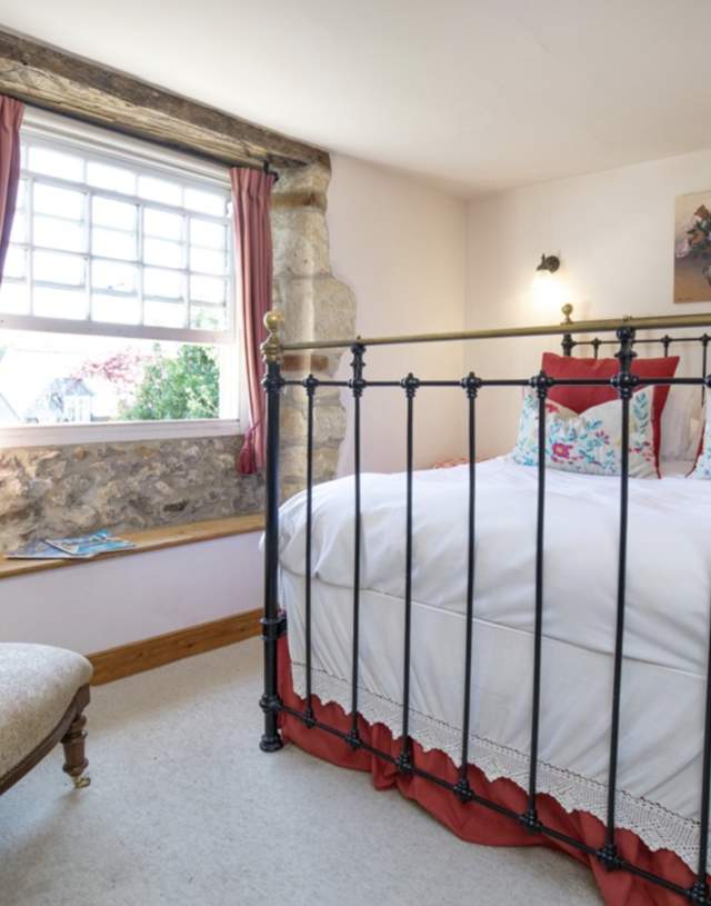Cosy bedroom with stone walls and rustic iron bed. Vibrant red and floral accents on pillows and decor. Sunlit window and elegant armchair. Warm ambience.