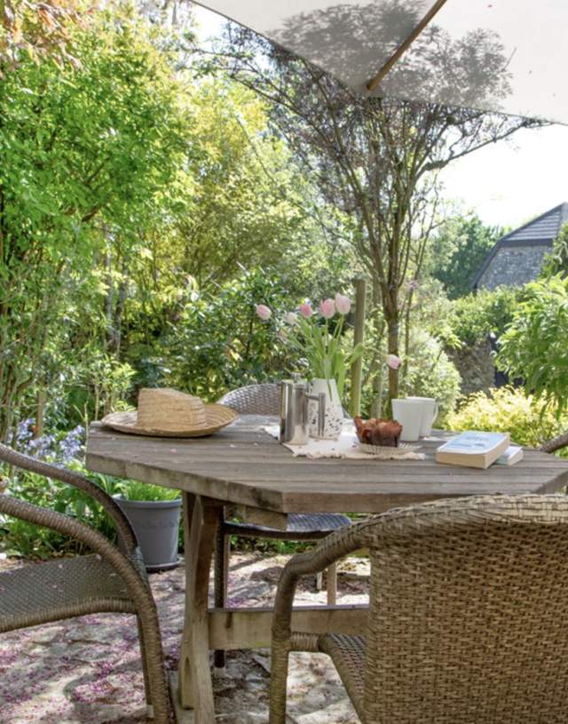 Outdoor garden scene with wicker chairs around a wooden table under a large umbrella. The table holds a book, flowers, and a hat, surrounded by lush greenery.
