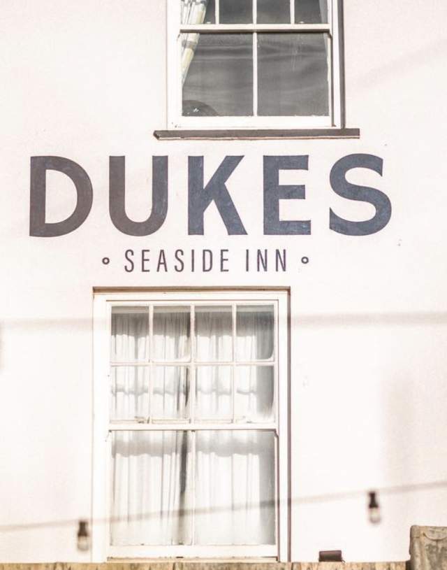 Facade of Dukes Seaside Inn with bold signage. The building features white walls and a curtained window. A relaxed, coastal ambiance is conveyed.