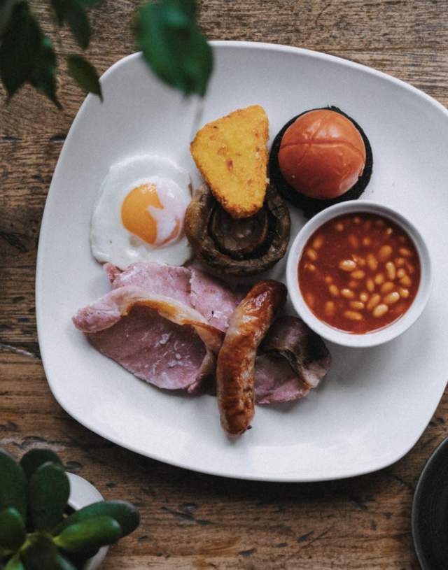 A hearty English breakfast with bacon, sausage, fried egg, baked beans, tomato, mushroom, hash brown, on a white plate. Tea, cutlery, and a newspaper on a wooden table. Cosy and inviting.