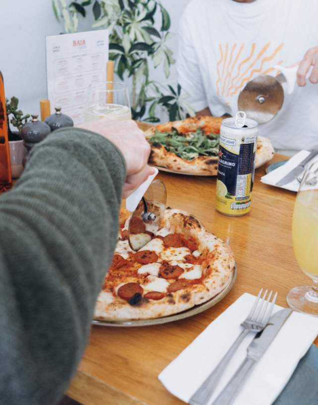 Two people at a table enjoy pizzas with vibrant toppings. Drinks and olive oil bottles adorn the table, creating a cosy and appetising atmosphere.