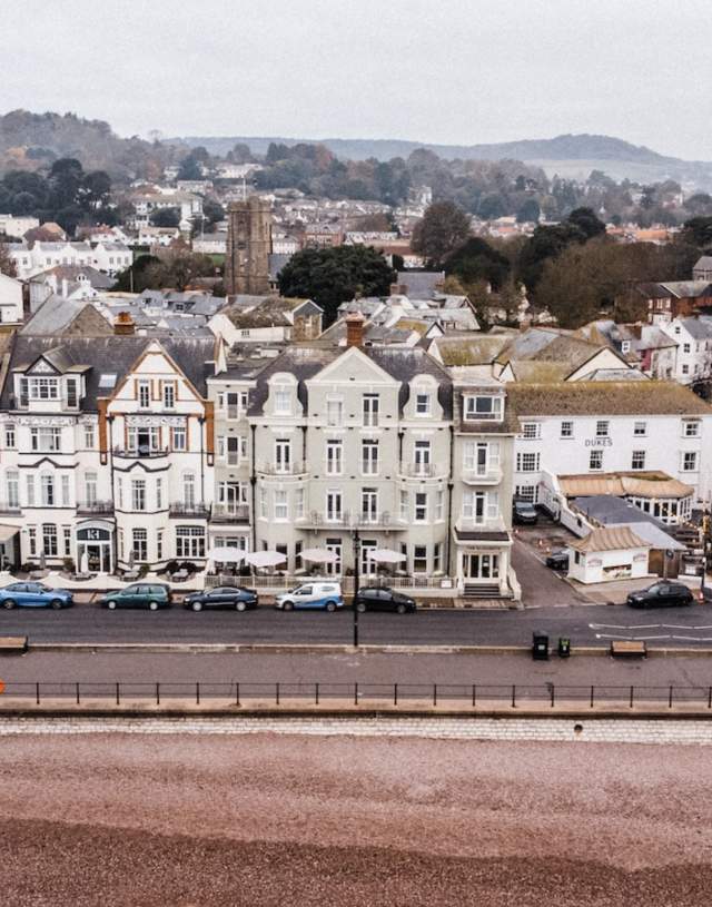 Aerial view of a coastal town with Victorian buildings along a promenade. Parked cars line the street, and hills are visible in the foggy background.