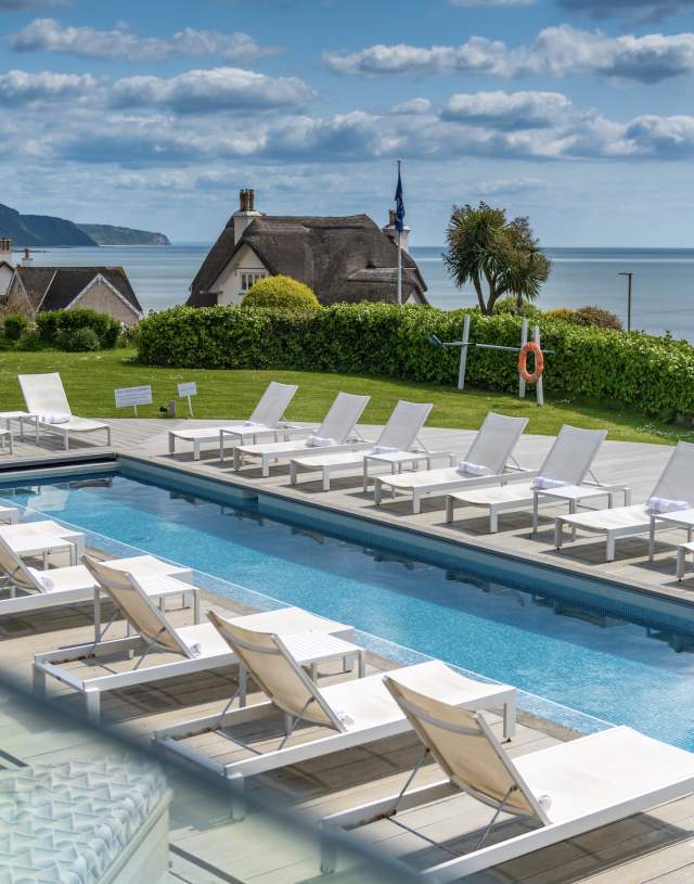 Outdoor pool area with white loungers, surrounded by green hedges in a coastal village. Thatched cottages and ocean view under a partly cloudy sky. Serene atmosphere.