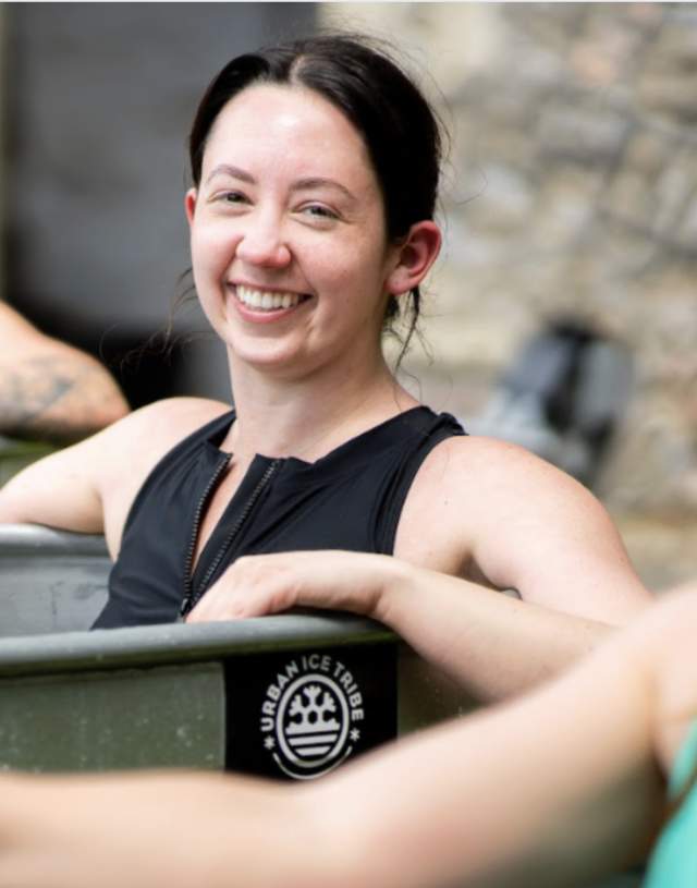 Three people smiling while sitting in ice baths outdoors. The central person wears a black swimsuit and appears joyful and relaxed. Blurred background.