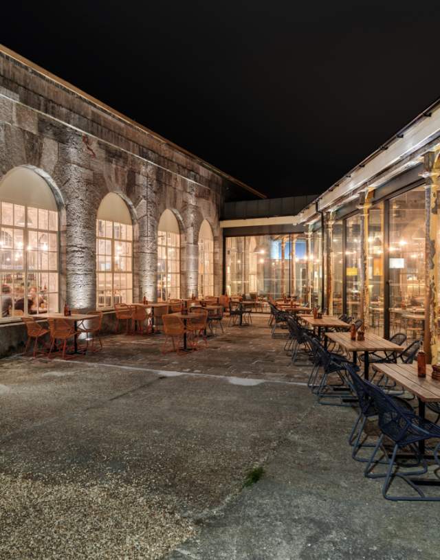 Outdoor dining area with empty tables and chairs under warm lights at night. Arched windows reveal a cosy, well-lit interior with a welcoming atmosphere.