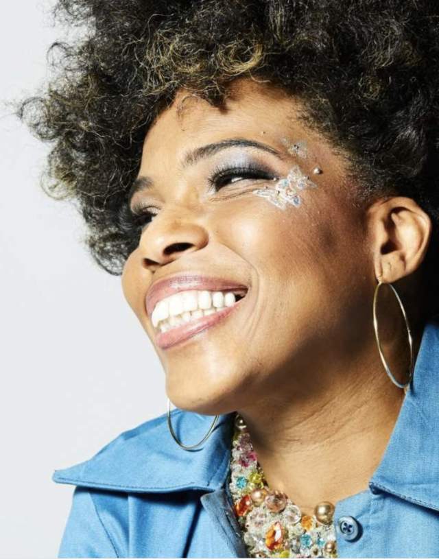Smiling woman with curly hair, wearing a blue top and hoop earrings, adorned with sparkly face jewels around her eyes, exudes joy and confidence.