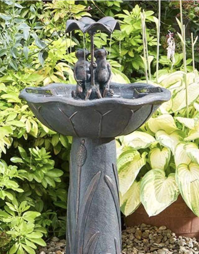 A stone fountain with two frog sculptures holding an umbrella, surrounded by lush garden foliage. It's a serene and whimsical garden scene.