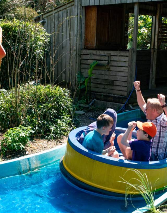 A group of people enjoy a log flume ride, passing by a large, whimsical orange flower sculpture. Lush greenery surrounds the area.