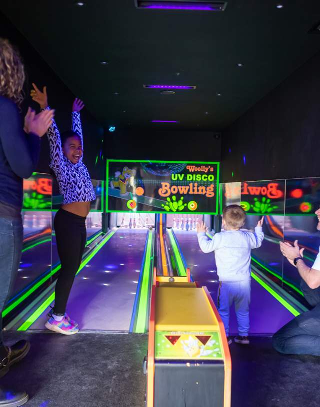 A child wearing a leopard print shirt raises their arms in excitement in a vibrant UV disco bowling alley. Nearby, two adults and another child clap and celebrate. The atmosphere is joyful and lively, with neon lights and colorful reflections.