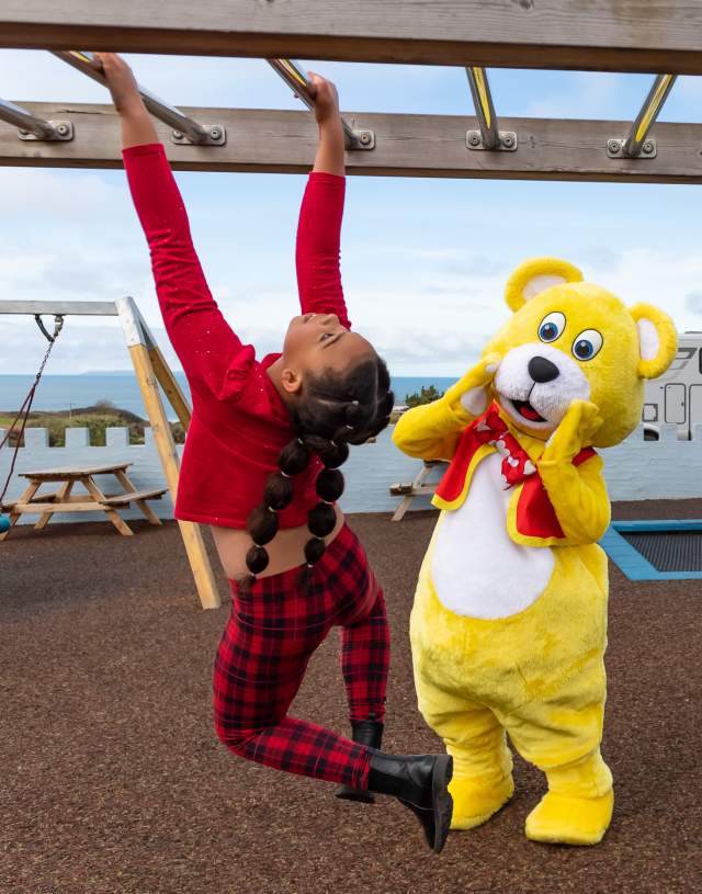 A child in red plaid pants playfully swings on monkey bars, while a person in a yellow bear costume stands nearby cheering. The background shows a playground with a sea view.
