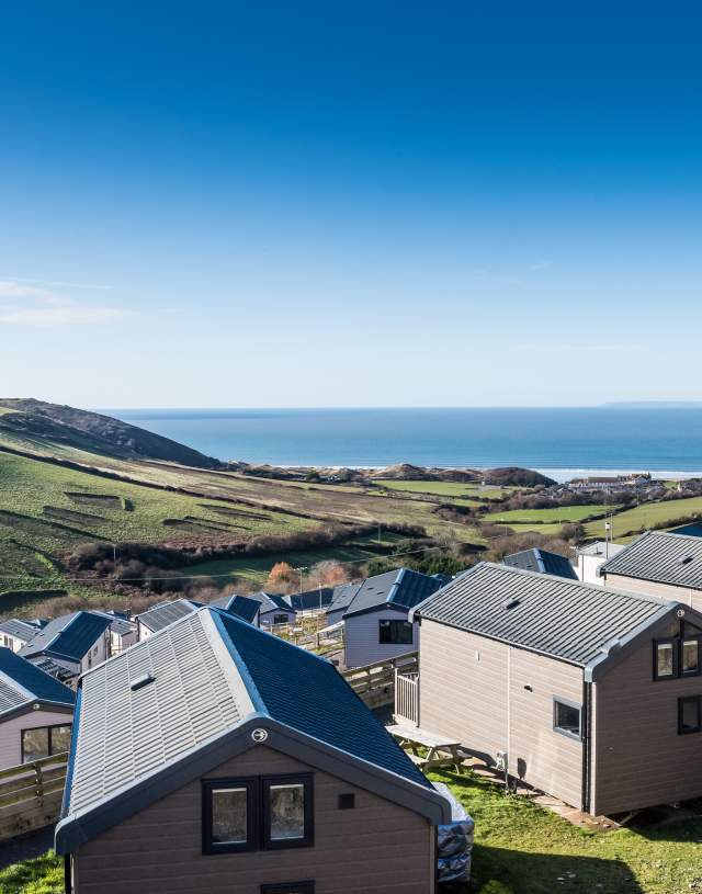Scenic view of a hillside with modern cabins overlooking the ocean. Green fields stretch toward the sea under a clear blue sky, conveying tranquillity.