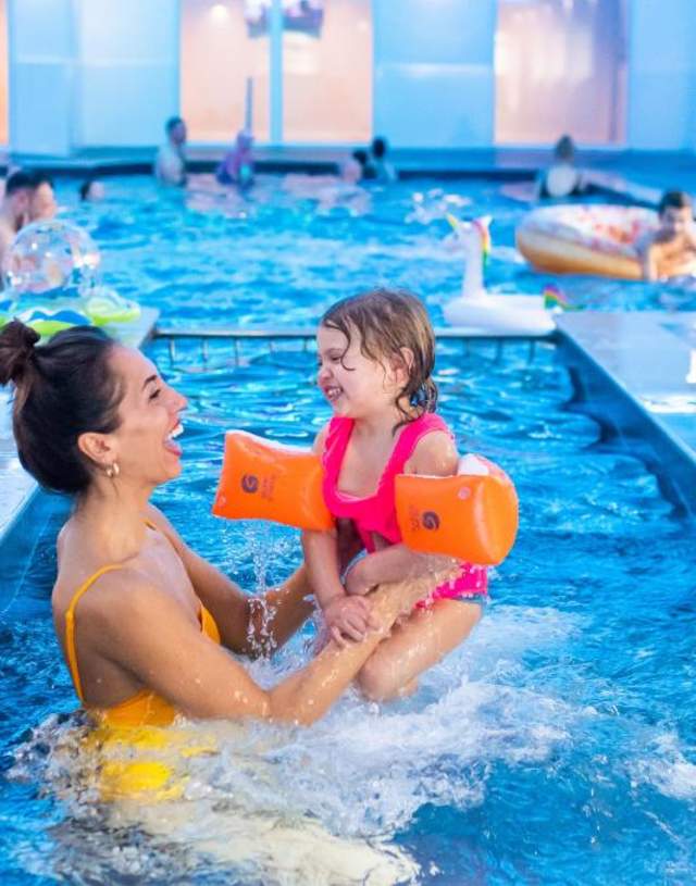 A woman in a yellow swimsuit joyfully holds a smiling child with orange floaties in a lively indoor pool. Other swimmers are visible in the background.