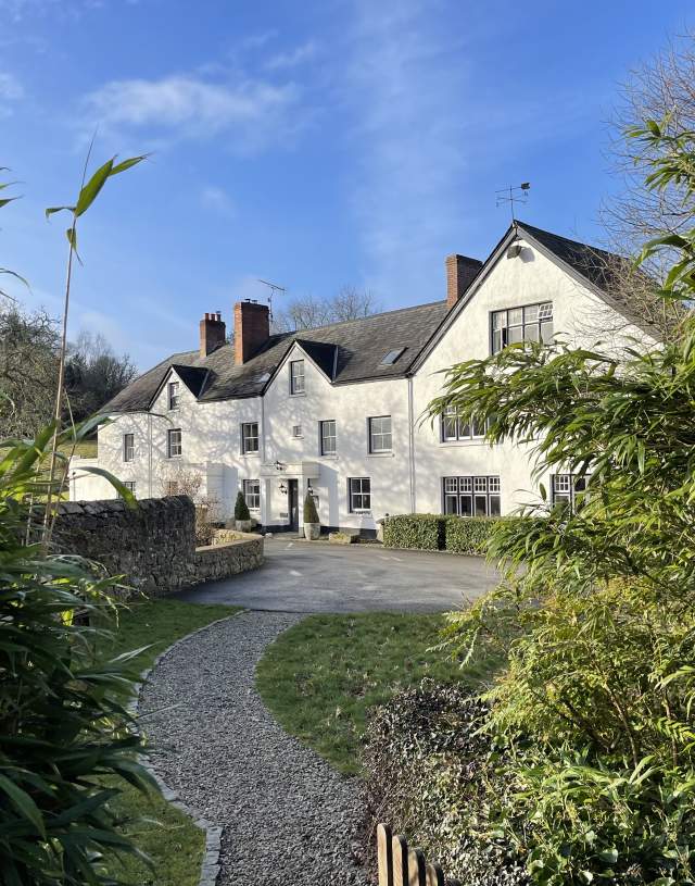 White country house with slate roof, surrounded by lush greenery and bamboo, under a clear blue sky. A gravel path leads to the entrance, creating a serene atmosphere.