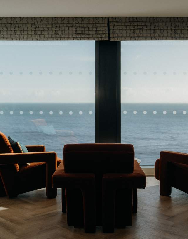 A tranquil room with three plush chairs facing a large window, offering a serene ocean view. A potted plant adds a touch of greenery to the cosy space.