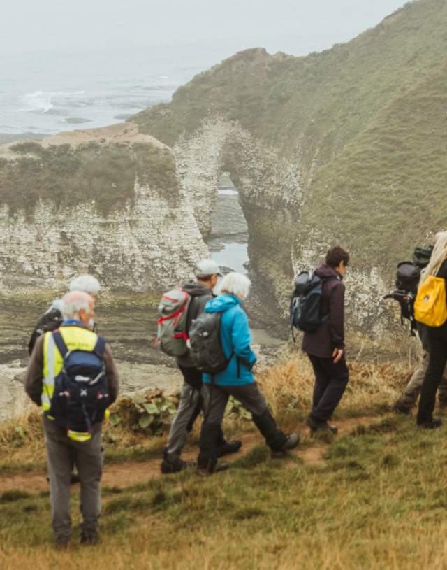 A group walking part of the KCIII England Coast Path in a long line, with the drinking dinosaur in the background on a foggy day.