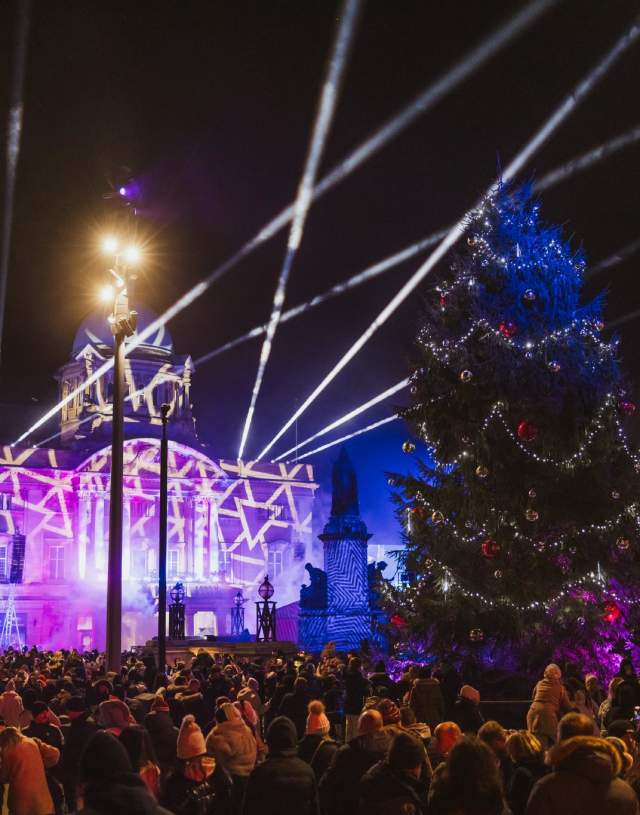 A large crowd gathers at night in a festive Queen Victoria Square, featuring a tall Christmas tree decorated with lights and ornaments, while an illuminated building with star-shaped light projections and beams shines in the background.