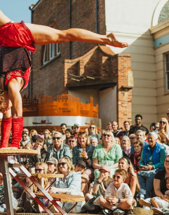 A woman in a red leotard is performing a hand stand on a raised platform with a crowd surrounding her.