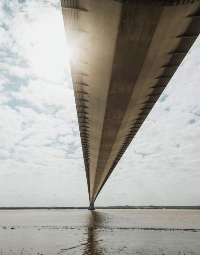 A view beneath the Humber bridge, with clouds either side and the foreshore just in front of the viewer.