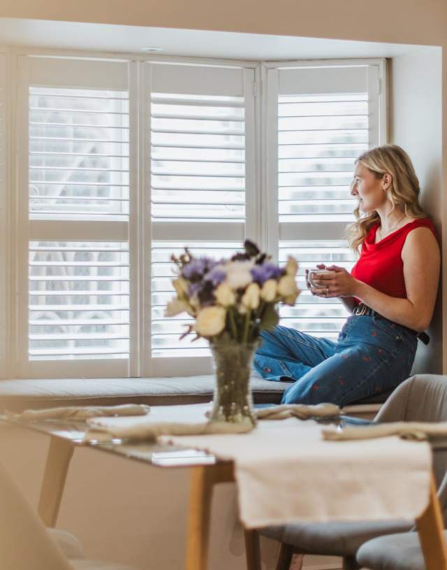 A girl in a red top sits on the inside of a set of bay windows, holding a mug and looking outside smiling. She has a table of flowers in front of her.