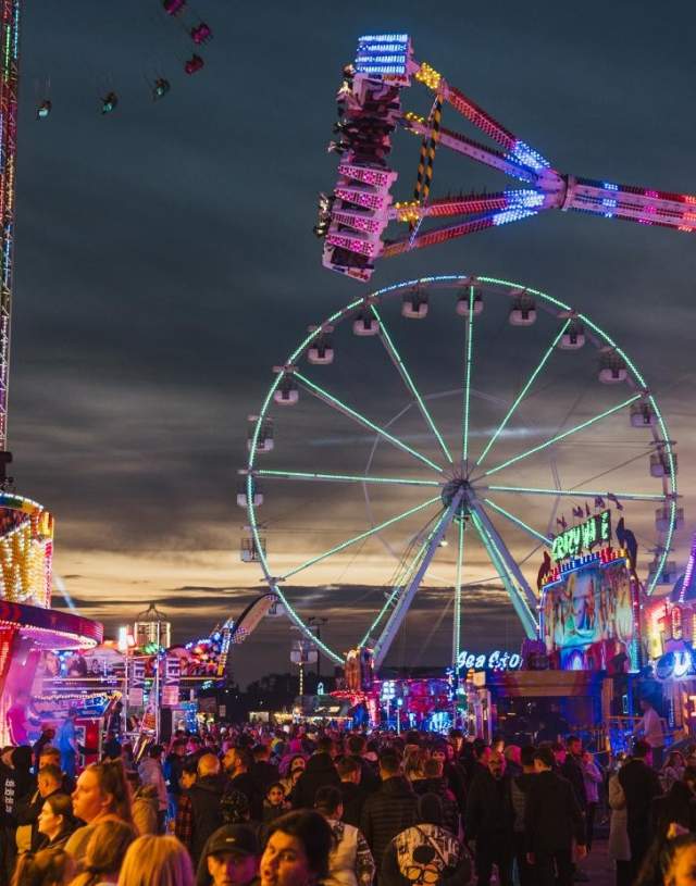 Showing crowds at the fair with bright neon lights and a ferries wheel in the background.