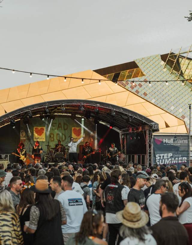 The crowd at Humber Street Sesh at sunset. The crowd are wearing summer clothes and are watching a band at the Hull Trains stage which is branded in the blue Hull Trains logo with hearts. Behind the stage, The Deep's iconic sloping arcitecture shines in the sunset.