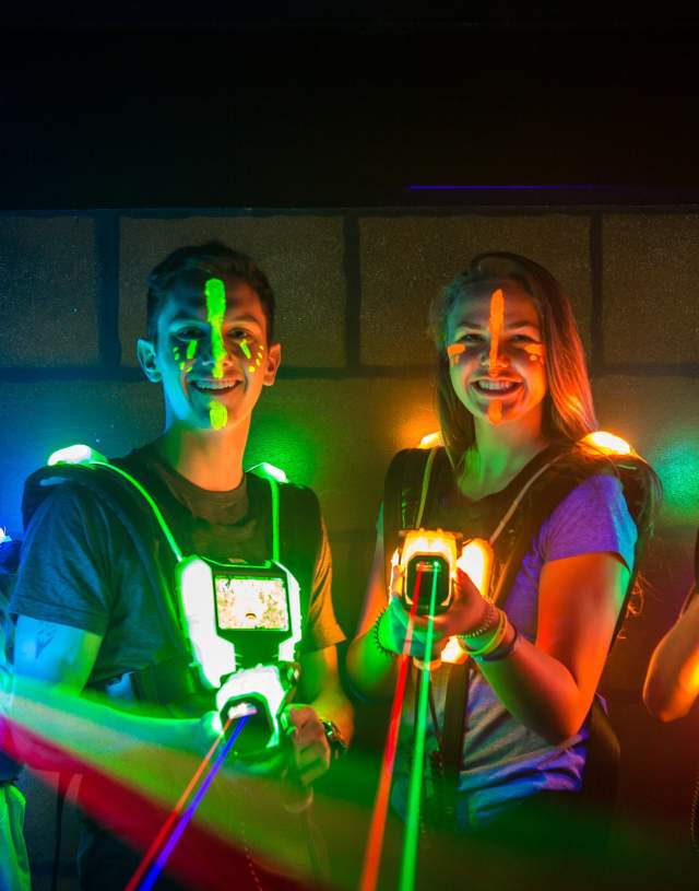 Four smiling young people stand side by side wearing glowing vests and holding laser tag guns, with colourful face paint and neon lights illuminating the dark room. Vibrant laser beams are visible shooting from their guns.