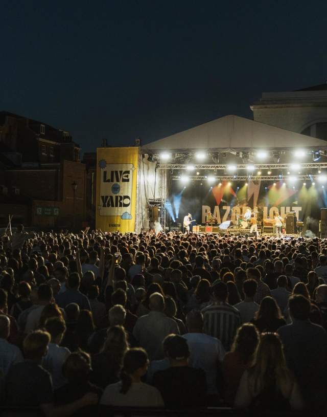 The stage at Live At The Yard at night. Razorlight are on stage infront of a banner emblazened with their logo. Bright lights are shining down on them and the crowd from the top of the pyramid shaped stage. Yellow banners flank both sides of the stage with Live At The Yard branding. In the foreground 100s of people are watching the band on stage.