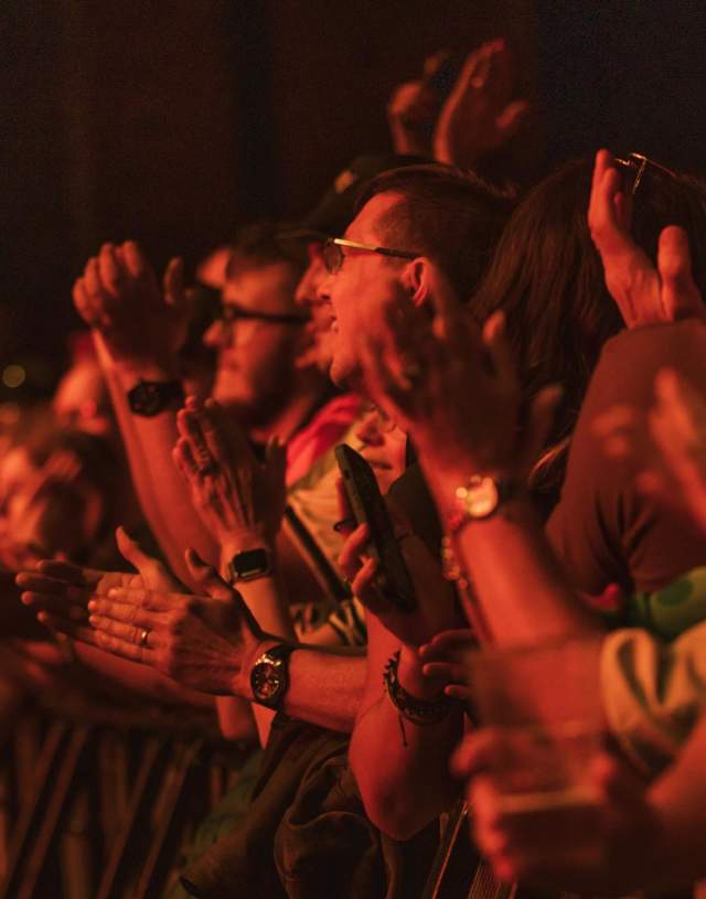 Conceert goers at Hull's Zebedee's Yard applauding and looking up at the stage. The lights from the stage are reflecting back at them washing the crowd with a red hue.