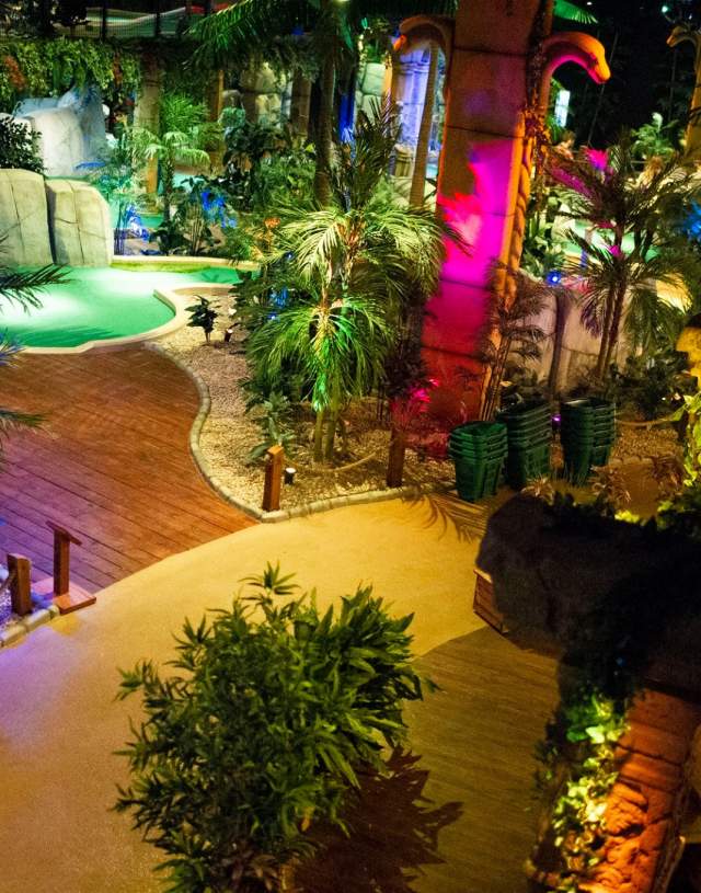 The brightly lit, Lost Coty Adventure Golf in Hull at night with winding wooden walkways, palm trees, rock formations, and a small green putting area, illuminated by colourful lights in shades of green, blue, purple, and pink.