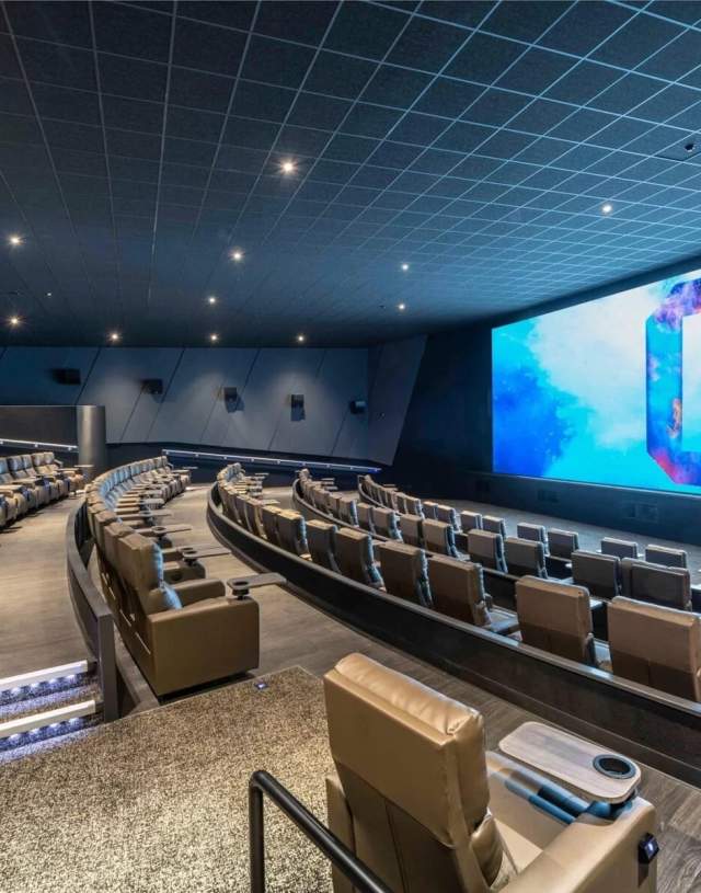 Modern movie theatre with several rows of large, reclining leather seats facing a wide screen displaying a colourful blue and white graphic. The theatre is spacious, dimly lit, and has cup holders and armrests on each seat.