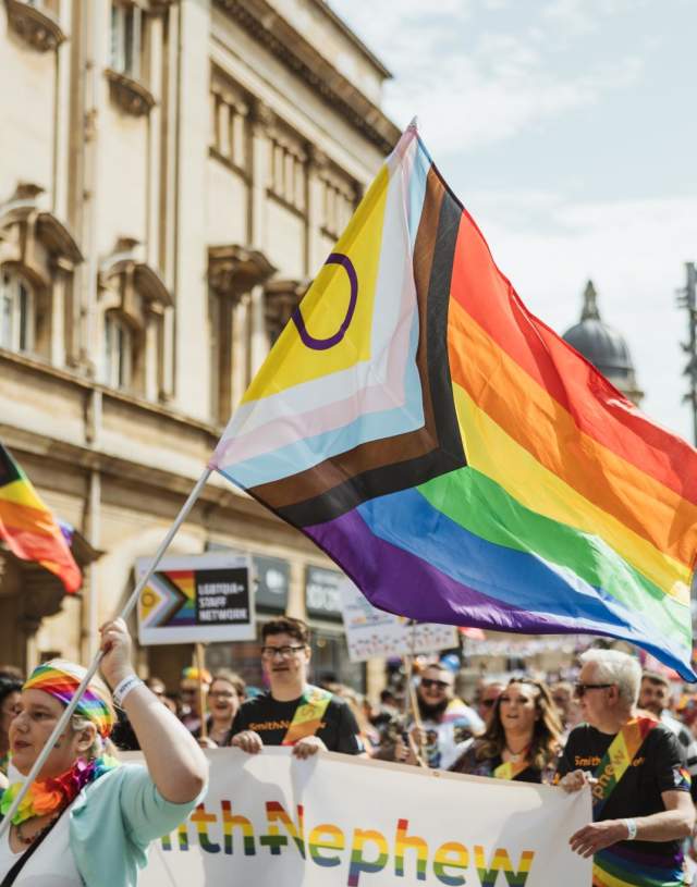Pride In Hull's parade walking down Carr Lane. The sun is shining, the crowd is smiling and dressed in shorts and t-shirts. A group of people holding a rainbow Smith+Nephew banner are in the foreground with a brightly dressed woman waving an Intersex Inclusive pride flag. Many other pride flags can be seen in the background.