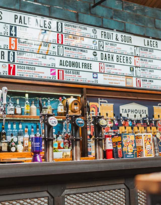 The interior of Taphouse on Humber Street in Hull, a wide assortment of colourful bottles sit behind a bar. Above sits a sign with a large variety of different drinks on offer.