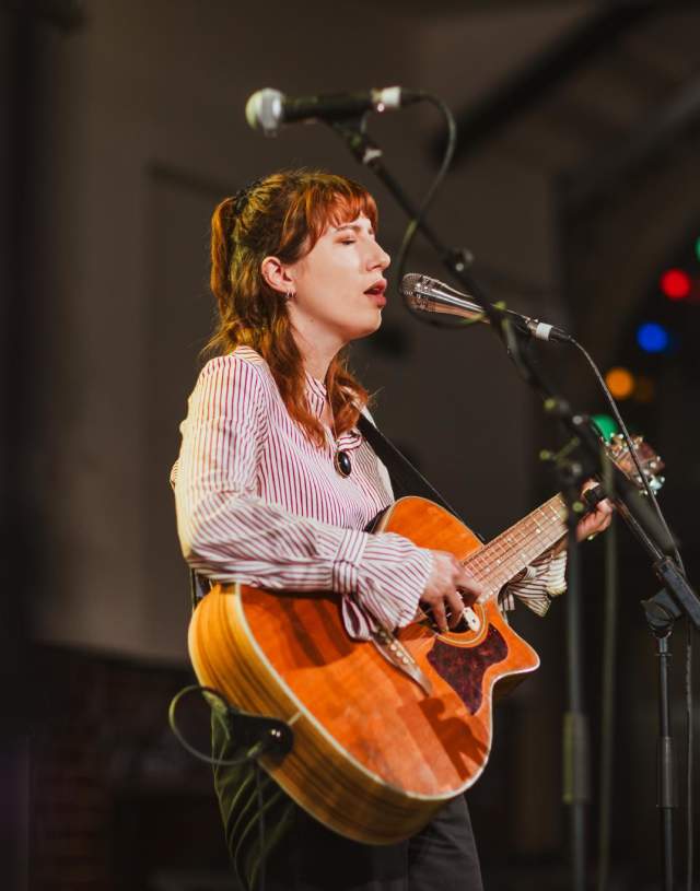 A woman plays an acoustic guitar onstage whilst singing into a microphone.
