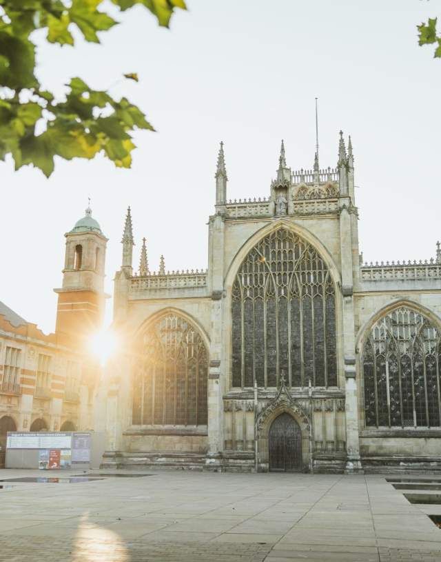 The exterior of Hull Minster at sunrise, a beautifully ornate church with a number of filials with the sun peeking through the edges of the buildings around it.