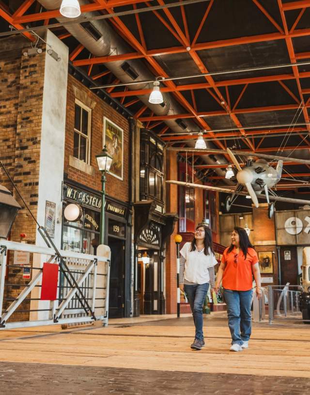 The main concourse of Streetlife Museum. The beamed ceilings are lied with bright lights and a propellor plane hangs from the rafter. Below is a replica 1950s high street. A vintage OXOsign is on the back wall with an retro van parked in front. Two women are walking underneath the plan across an old train cross towards the camera. They are looking up at the exhibits and are wearing t-shirts and jeans.