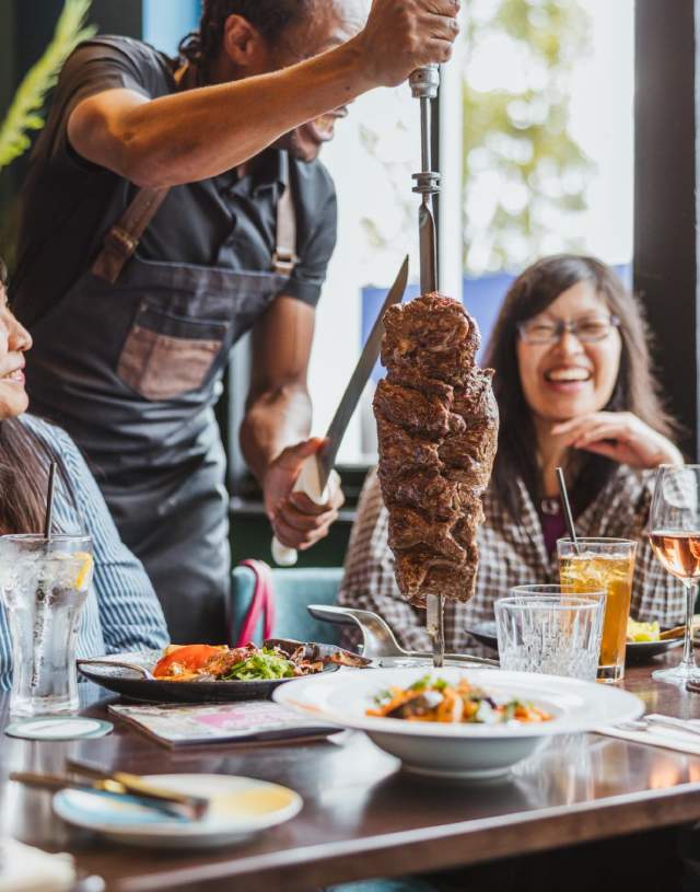 Two women sat at a table covered in food being served by a waiter with a large kebab of meat on a skewer.
