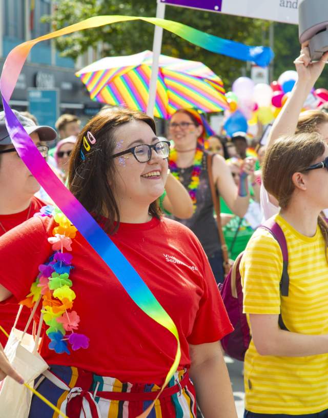 Woman smiling waving rainbow ribbon in Southampton Pride march through Above Bar Street