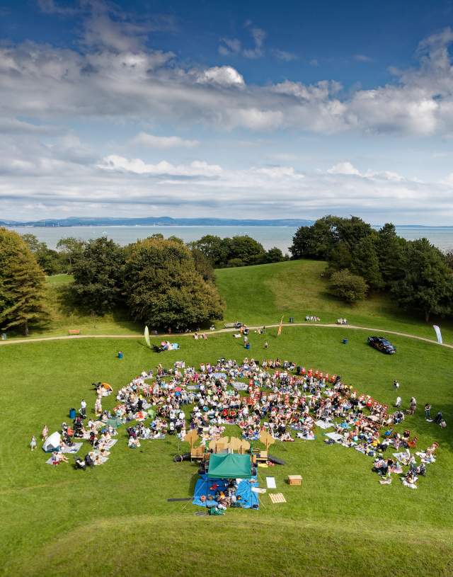 aerial photo of crowd watching an outdoor theatre performance with swansea bay and mumbles in the background